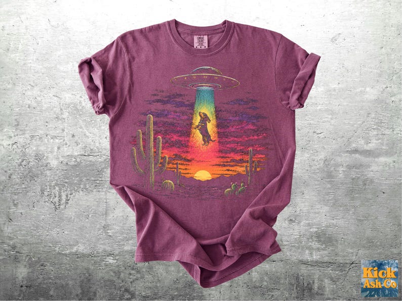 May include: A maroon short-sleeve t-shirt featuring a graphic of a dachshund being abducted by a UFO in a desert landscape at sunset. The design includes a bright beam of light, saguaro cacti, and a vibrant sky with purple and orange hues.
