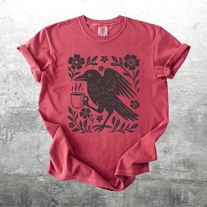 May include: Red t-shirt featuring a black illustration of a crow holding a steaming coffee cup, surrounded by flowers and leaves. The shirt has rolled-up sleeves and a crew neck. The design is centered on the front of the shirt.