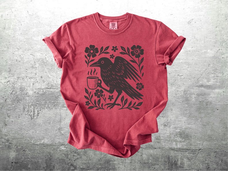 May include: Red t-shirt featuring a black illustration of a crow holding a steaming coffee cup, surrounded by flowers and leaves. The shirt has rolled-up sleeves and a crew neck. The design is centered on the front of the shirt.