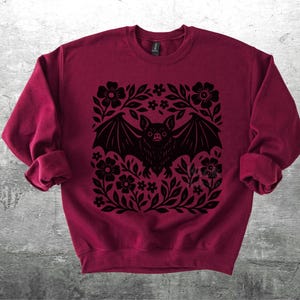 May include: A burgundy sweatshirt featuring a black bat design surrounded by flowers and leaves. The bat has outstretched wings and is centered within the floral arrangement. The sweatshirt has long sleeves and a crew neck.