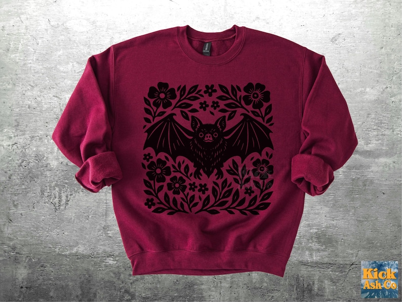 May include: A burgundy sweatshirt featuring a black bat design surrounded by flowers and leaves. The bat has outstretched wings and is centered within the floral arrangement. The sweatshirt has long sleeves and a crew neck.