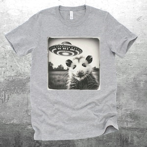 May include: A gray t-shirt with a black and white image of a possum looking up at a flying saucer. The image is in a vintage style.