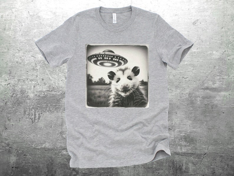 May include: A gray t-shirt with a black and white image of a possum looking up at a flying saucer. The image is in a vintage style.