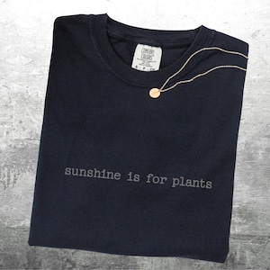 May include: A navy blue t-shirt with the text "sunshine is for plants" printed in grey. A gold necklace with a circular pendant is draped on the shirt. The shirt has a Comfort Colors label.