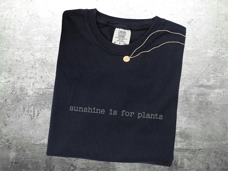 May include: A navy blue t-shirt with the text "sunshine is for plants" printed in grey. A gold necklace with a circular pendant is draped on the shirt. The shirt has a Comfort Colors label.