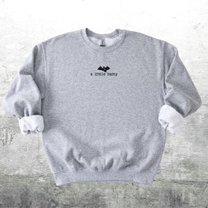 May include: A heather gray sweatshirt with the phrase "a little batty" and a bat graphic embroidered in black. The sleeves are cuffed with white fabric. The sweatshirt is a casual, comfortable garment.