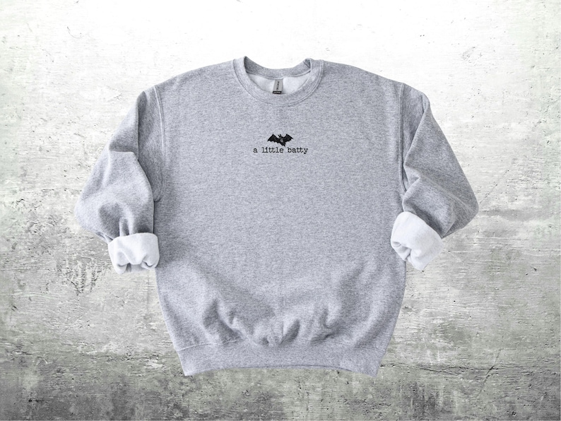 May include: A heather gray sweatshirt with the phrase "a little batty" and a bat graphic embroidered in black. The sleeves are cuffed with white fabric. The sweatshirt is a casual, comfortable garment.