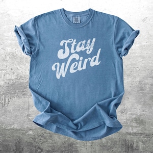 K&ouml;nnte beinhalten: Ein blaues T-Shirt mit dem wei&szlig;en Schriftzug "Stay Weird" in verschn&ouml;rkelter Schrift. Das Shirt besteht aus einem weichen Material und hat hochgekrempelte &Auml;rmel. Das Shirt wird auf einer grauen, strukturierten Oberfl&auml;che pr&auml;sentiert.