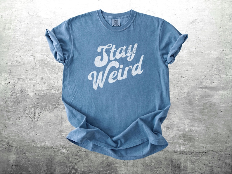 K&ouml;nnte beinhalten: Ein blaues T-Shirt mit dem wei&szlig;en Schriftzug "Stay Weird" in verschn&ouml;rkelter Schrift. Das Shirt besteht aus einem weichen Material und hat hochgekrempelte &Auml;rmel. Das Shirt wird auf einer grauen, strukturierten Oberfl&auml;che pr&auml;sentiert.
