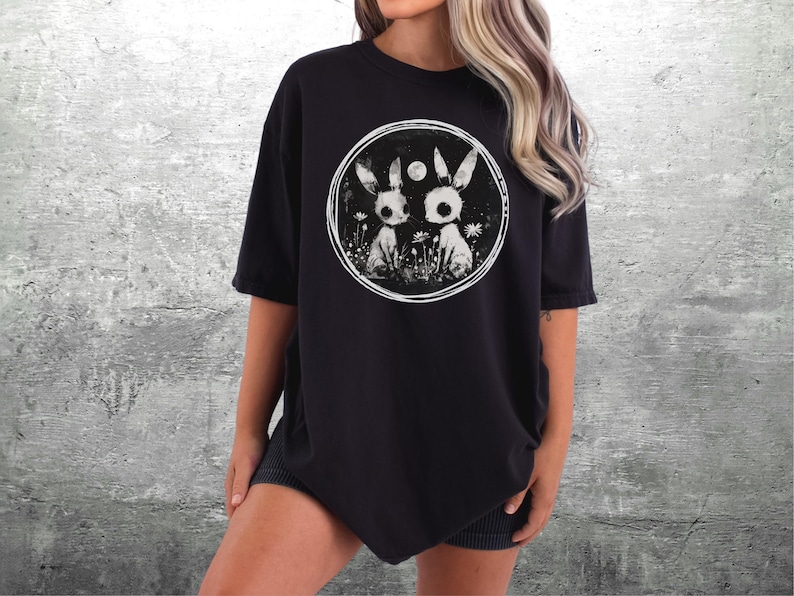 Op de afbeelding: Zwart oversized T-shirt met een grafische print van twee witte konijnen met zwarte ogen in een cirkel. De konijnen zijn omringd door witte bloemen en een halve maan.