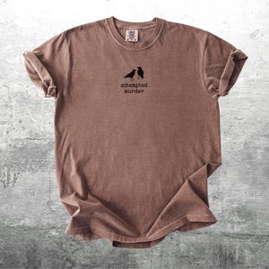 May include: Brown t-shirt with rolled sleeves. The shirt features a small graphic of two black birds and the text "attempted murder" in a simple font. The shirt is displayed on a textured gray surface.