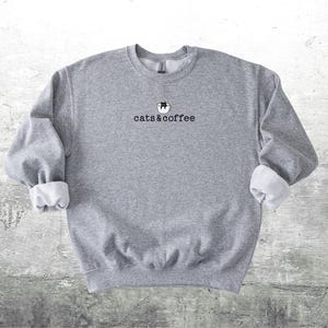 May include: A gray sweatshirt with the text "cats & coffee" in black. The sweatshirt has a small graphic of a cat in a coffee cup above the text. The sleeves are cuffed, revealing a white lining.