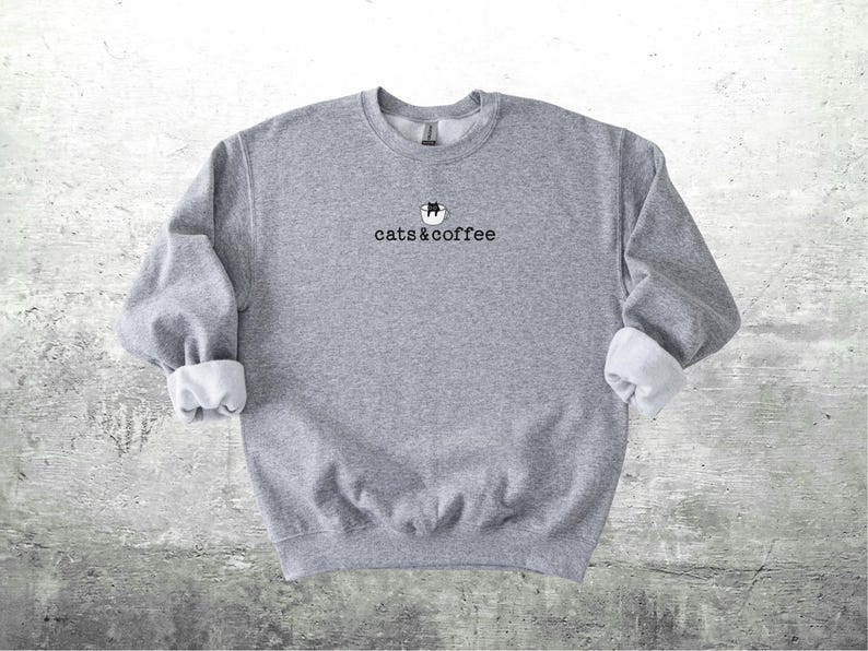 May include: A gray sweatshirt with the text "cats & coffee" in black. The sweatshirt has a small graphic of a cat in a coffee cup above the text. The sleeves are cuffed, revealing a white lining.