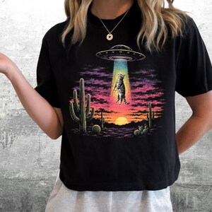 May include: A black short-sleeve t-shirt featuring a graphic print of a tabby cat being abducted by a UFO in a desert landscape at sunset. The design includes saguaro cacti, a glowing blue tractor beam, and a vibrant purple and orange sky.