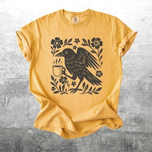 May include: Mustard yellow t-shirt featuring a black graphic of a crow holding a coffee cup, surrounded by flowers and leaves. The shirt has a crew neck and short sleeves. The design is centered on the front of the shirt.