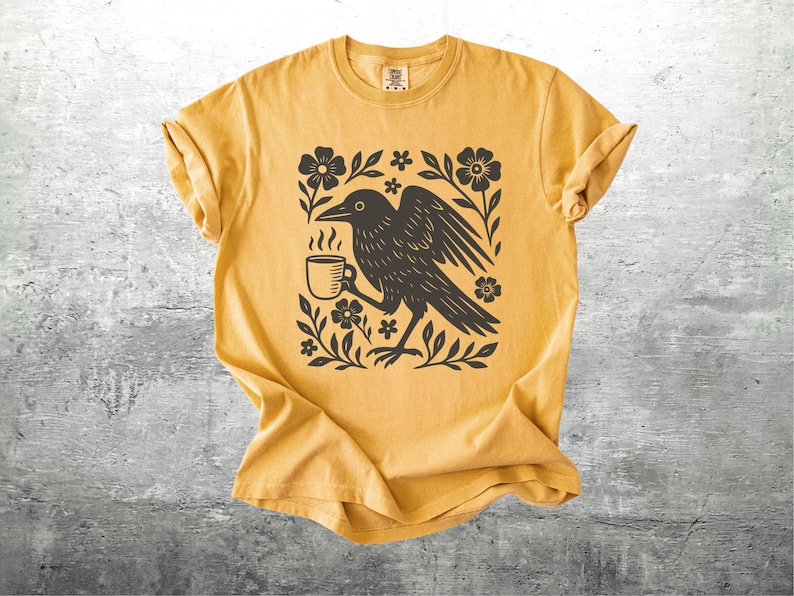 May include: Mustard yellow t-shirt featuring a black graphic of a crow holding a coffee cup, surrounded by flowers and leaves. The shirt has a crew neck and short sleeves. The design is centered on the front of the shirt.