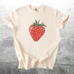 May include: Cream-colored t-shirt featuring a large, distressed red strawberry graphic with white seeds and green stem. The shirt is made of soft material and has rolled-up sleeves.