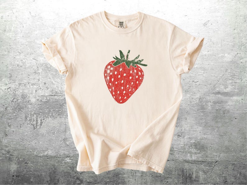 May include: Cream-colored t-shirt featuring a large, distressed red strawberry graphic with white seeds and green stem. The shirt is made of soft material and has rolled-up sleeves.