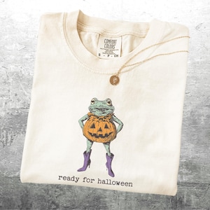 May include: Cream-colored t-shirt with a graphic of a frog in a pumpkin costume and purple boots. The text "ready for halloween" is printed below the frog. A gold necklace with a pendant is draped over the shirt.