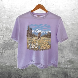 Puede incluir: Camiseta de color lila claro con un gr&aacute;fico de un Bigfoot en un campo con monta&ntilde;as y &aacute;rboles. El Bigfoot es marr&oacute;n y las monta&ntilde;as son azules y blancas. La camiseta est&aacute; colgada en una percha de madera.