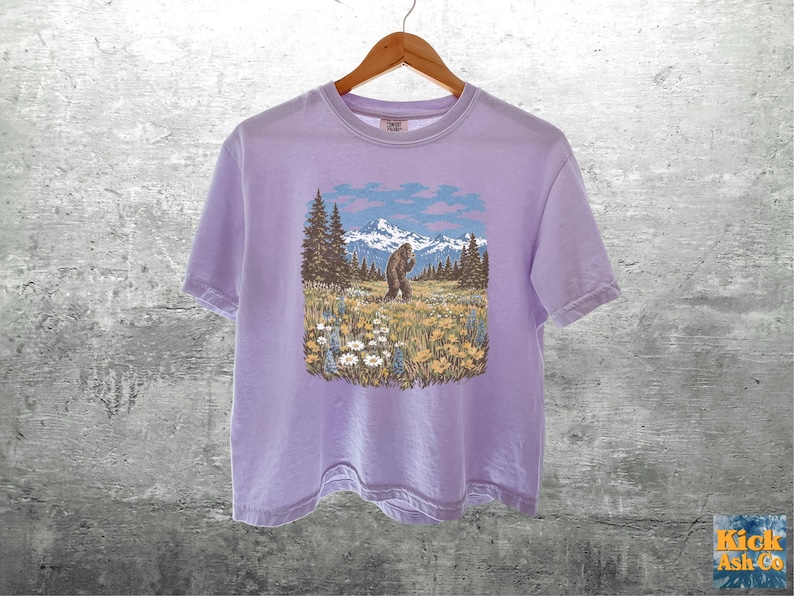 Puede incluir: Camiseta de color lila claro con un gr&aacute;fico de un Bigfoot en un campo con monta&ntilde;as y &aacute;rboles. El Bigfoot es marr&oacute;n y las monta&ntilde;as son azules y blancas. La camiseta est&aacute; colgada en una percha de madera.