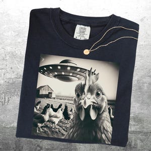 May include: Navy blue t-shirt featuring a black and white graphic of a chicken looking up at a UFO. The image includes a barn and other chickens in the background. A gold necklace with a small pendant is draped on the shirt.