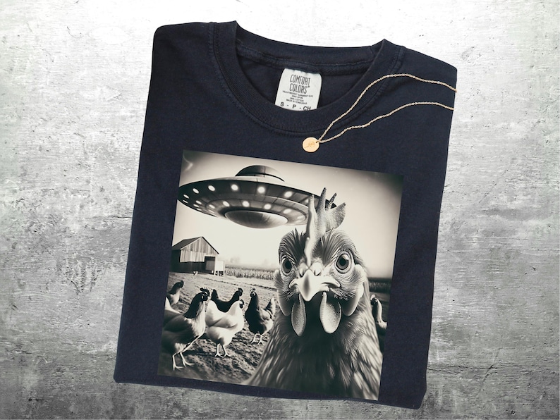 May include: Navy blue t-shirt featuring a black and white graphic of a chicken looking up at a UFO. The image includes a barn and other chickens in the background. A gold necklace with a small pendant is draped on the shirt.