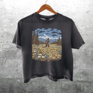 Puede incluir: Camiseta gris oscuro con un gr&aacute;fico de Bigfoot en un paisaje monta&ntilde;oso. El dise&ntilde;o incluye una figura de Bigfoot, &aacute;rboles, flores, monta&ntilde;as y un cielo azul con nubes. La camiseta est&aacute; colgada en una percha de madera.