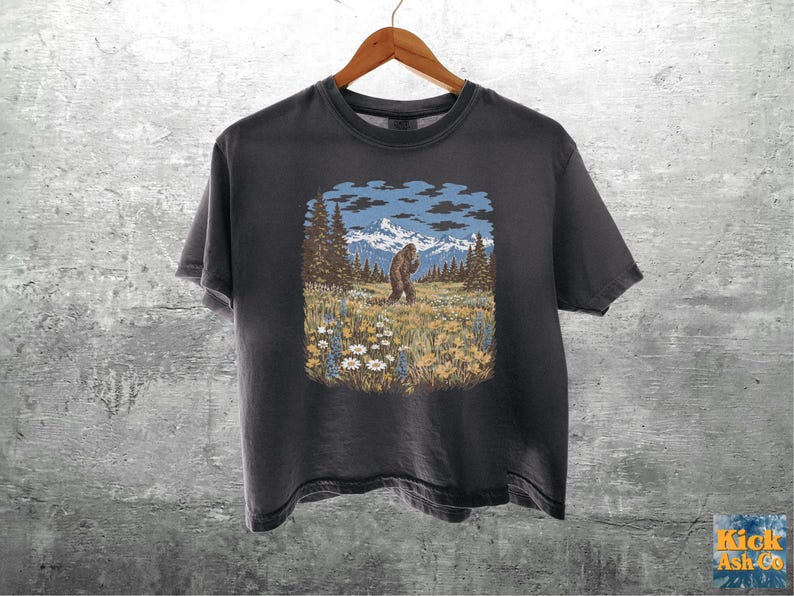 Puede incluir: Camiseta gris oscuro con un gr&aacute;fico de Bigfoot en un paisaje monta&ntilde;oso. El dise&ntilde;o incluye una figura de Bigfoot, &aacute;rboles, flores, monta&ntilde;as y un cielo azul con nubes. La camiseta est&aacute; colgada en una percha de madera.