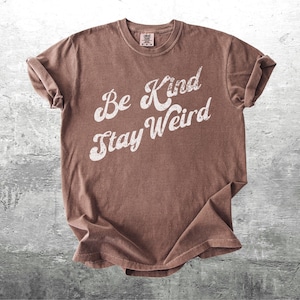 May include: A brown t-shirt with the words "Be Kind Stay Weird" in white, stylized lettering. The shirt has a vintage, slightly faded look and rolled-up sleeves. The tee is made of soft material.