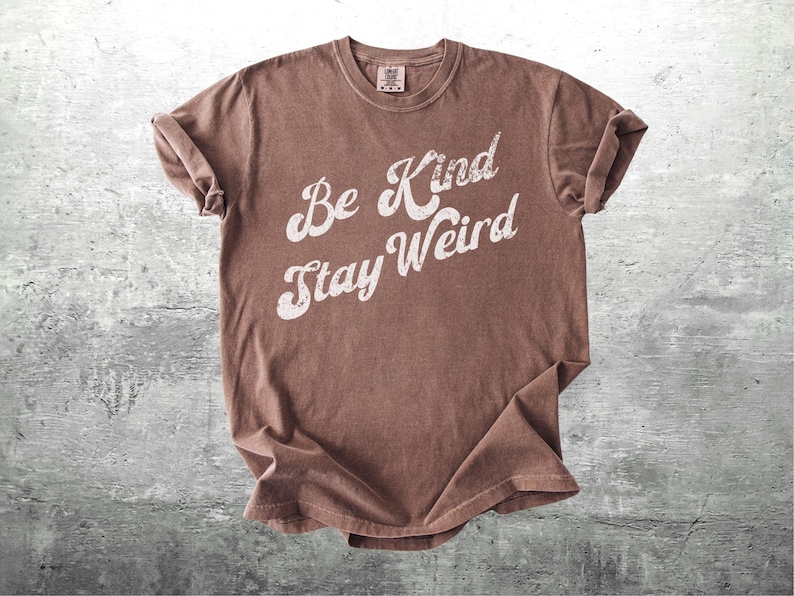 May include: A brown t-shirt with the words "Be Kind Stay Weird" in white, stylized lettering. The shirt has a vintage, slightly faded look and rolled-up sleeves. The tee is made of soft material.