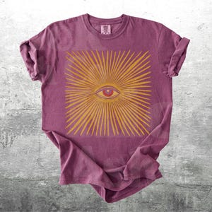 May include: A maroon t-shirt featuring a gold-colored eye design with radiating lines. The eye has a red iris and is centered within a square. The shirt has rolled-up sleeves and a crew neck.