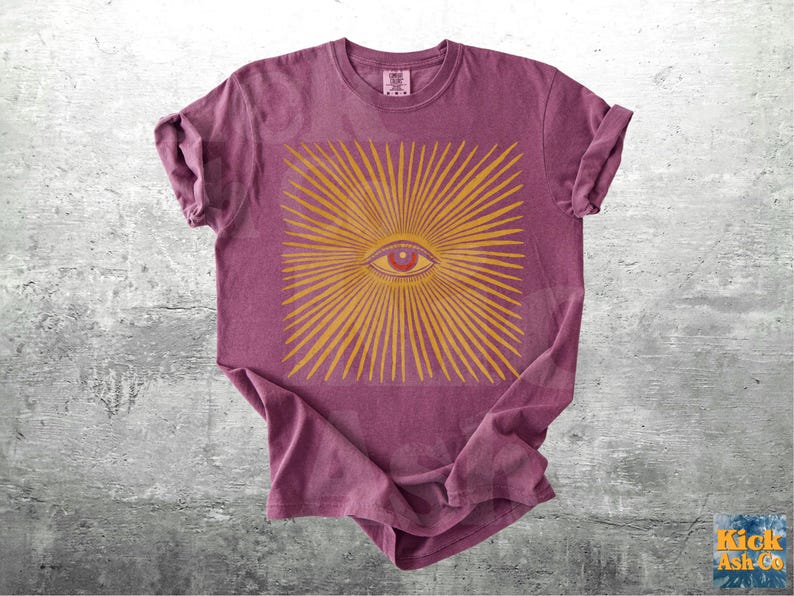 May include: A maroon t-shirt featuring a gold-colored eye design with radiating lines. The eye has a red iris and is centered within a square. The shirt has rolled-up sleeves and a crew neck.