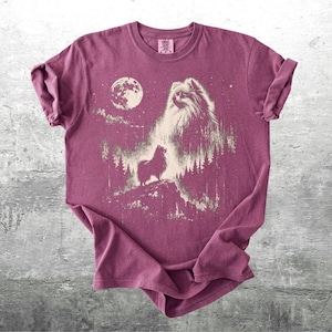 May include: A maroon t-shirt featuring a graphic of a dog with a full moon and forest scene. The dog is depicted in a detailed, artistic style. The shirt has rolled-up sleeves and is displayed on a gray surface.