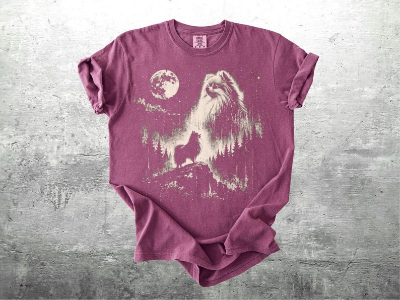 May include: A maroon t-shirt featuring a graphic of a dog with a full moon and forest scene. The dog is depicted in a detailed, artistic style. The shirt has rolled-up sleeves and is displayed on a gray surface.