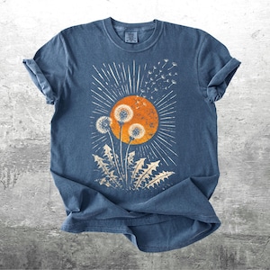 May include: A blue t-shirt featuring a dandelion design. The graphic includes a large orange sun with radiating lines, three dandelion seed heads, and dandelion leaves. The design is distressed for a vintage look.