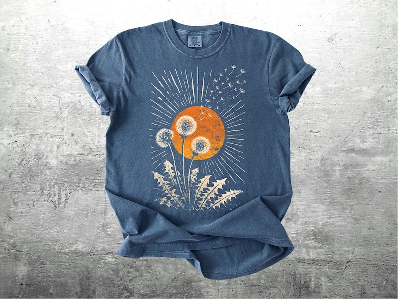 May include: A blue t-shirt featuring a dandelion design. The graphic includes a large orange sun with radiating lines, three dandelion seed heads, and dandelion leaves. The design is distressed for a vintage look.