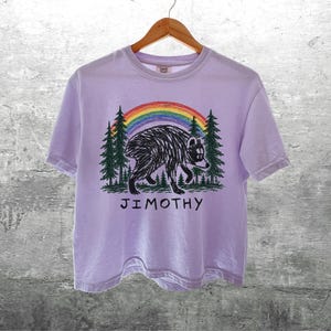 May include: A light purple short-sleeve t-shirt featuring a graphic of a black bear walking through a forest of green pine trees under a colorful rainbow. The text JIMOTHY is printed in black capital letters below the bear illustration.