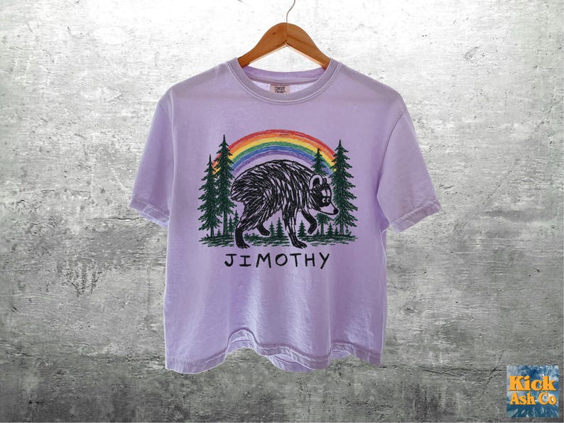 May include: A light purple short-sleeve t-shirt featuring a graphic of a black bear walking through a forest of green pine trees under a colorful rainbow. The text JIMOTHY is printed in black capital letters below the bear illustration.