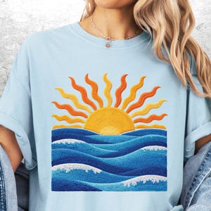 May include: Light blue t-shirt featuring an embroidered design of a yellow sun with orange rays rising above blue ocean waves. The waves have white foam details. The shirt has short sleeves and a crew neck.