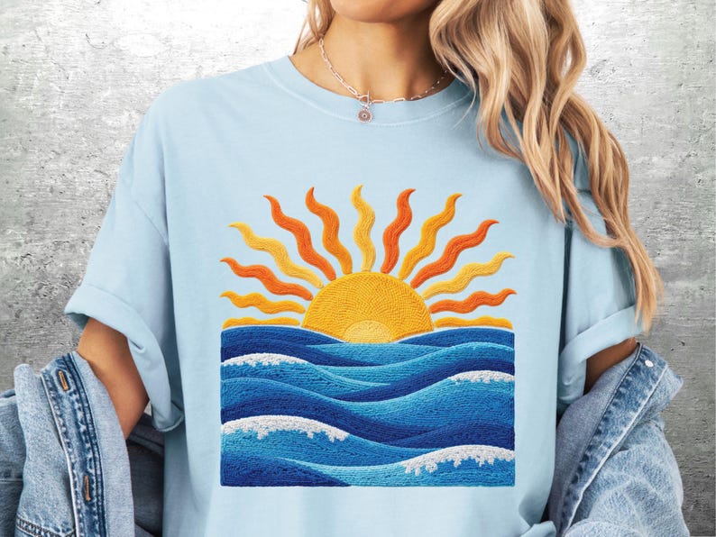 May include: Light blue t-shirt featuring an embroidered design of a yellow sun with orange rays rising above blue ocean waves. The waves have white foam details. The shirt has short sleeves and a crew neck.
