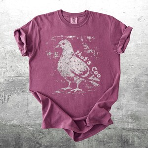 May include: A maroon t-shirt with a distressed white graphic of a pigeon and the words "that's Coo". The shirt is folded, showing the rolled-up sleeves and the tag on the collar.