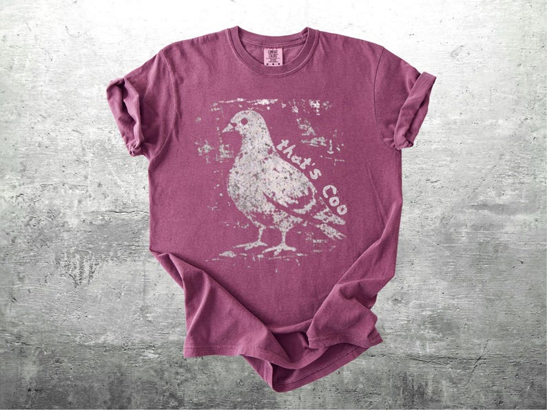 May include: A maroon t-shirt with a distressed white graphic of a pigeon and the words "that's Coo". The shirt is folded, showing the rolled-up sleeves and the tag on the collar.