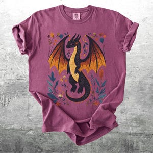 May include: A maroon t-shirt with a black and orange dragon illustration. The dragon is surrounded by floral designs in shades of yellow, orange, and green.