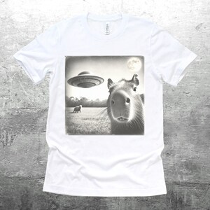 K&ouml;nnte beinhalten: Ein wei&szlig;es T-Shirt mit einem Schwarzwei&szlig;bild eines Capybaras, das in die Kamera schaut. Das Bild zeigt auch eine fliegende Untertasse im Hintergrund.