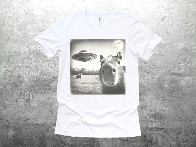 K&ouml;nnte beinhalten: Ein wei&szlig;es T-Shirt mit einem Schwarzwei&szlig;bild eines Capybaras, das in die Kamera schaut. Das Bild zeigt auch eine fliegende Untertasse im Hintergrund.