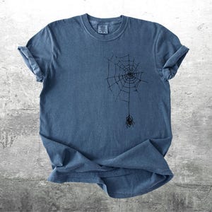 May include: Blue t-shirt with a black spider web design and a spider hanging from the web. The shirt has rolled-up sleeves and a crew neck. The fabric has a slightly faded appearance.