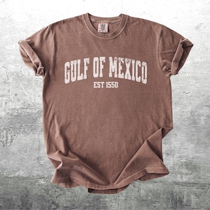 May include: A brown t-shirt with the text "GULF OF MEXICO EST 1550" printed in white.