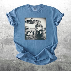 May include: A blue t-shirt featuring a black and white image of a chicken looking at a UFO. The image is centered on the shirt and shows a barn and other chickens in the background. The shirt is made of a soft material.