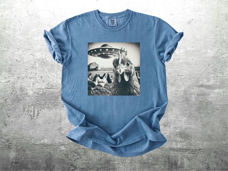 May include: A blue t-shirt featuring a black and white image of a chicken looking at a UFO. The image is centered on the shirt and shows a barn and other chickens in the background. The shirt is made of a soft material.